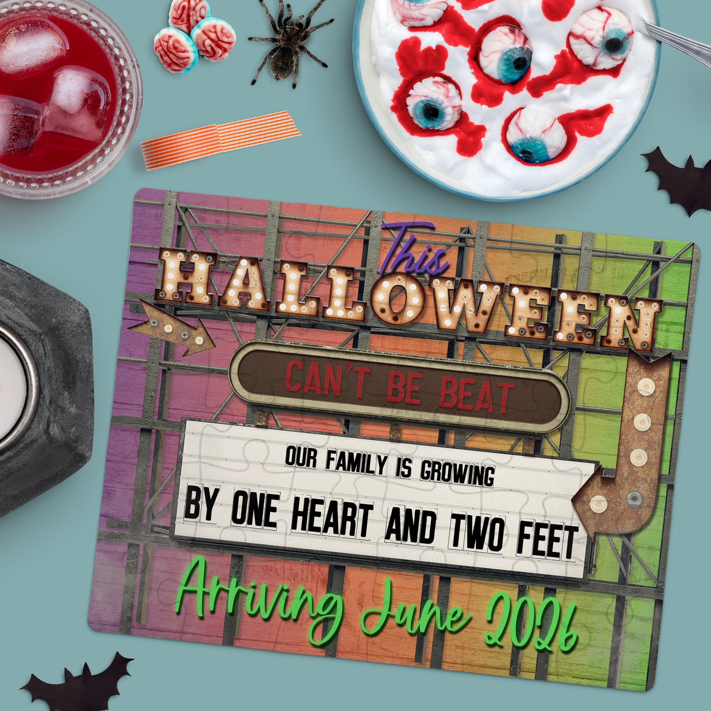 A Halloween-themed flat lay with a sign announcing a pregnancy: "Our family is growing by one heart and two feet. Arriving June 2026." Surrounded by spooky decorations and snacks.
