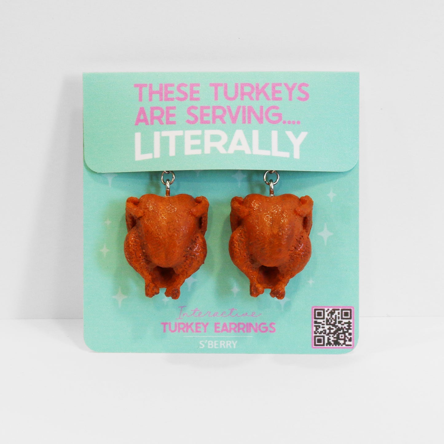 A pair of earrings shaped like roasted turkeys displayed on a card with the text "THESE TURKEYS ARE SERVING... LITERALLY.