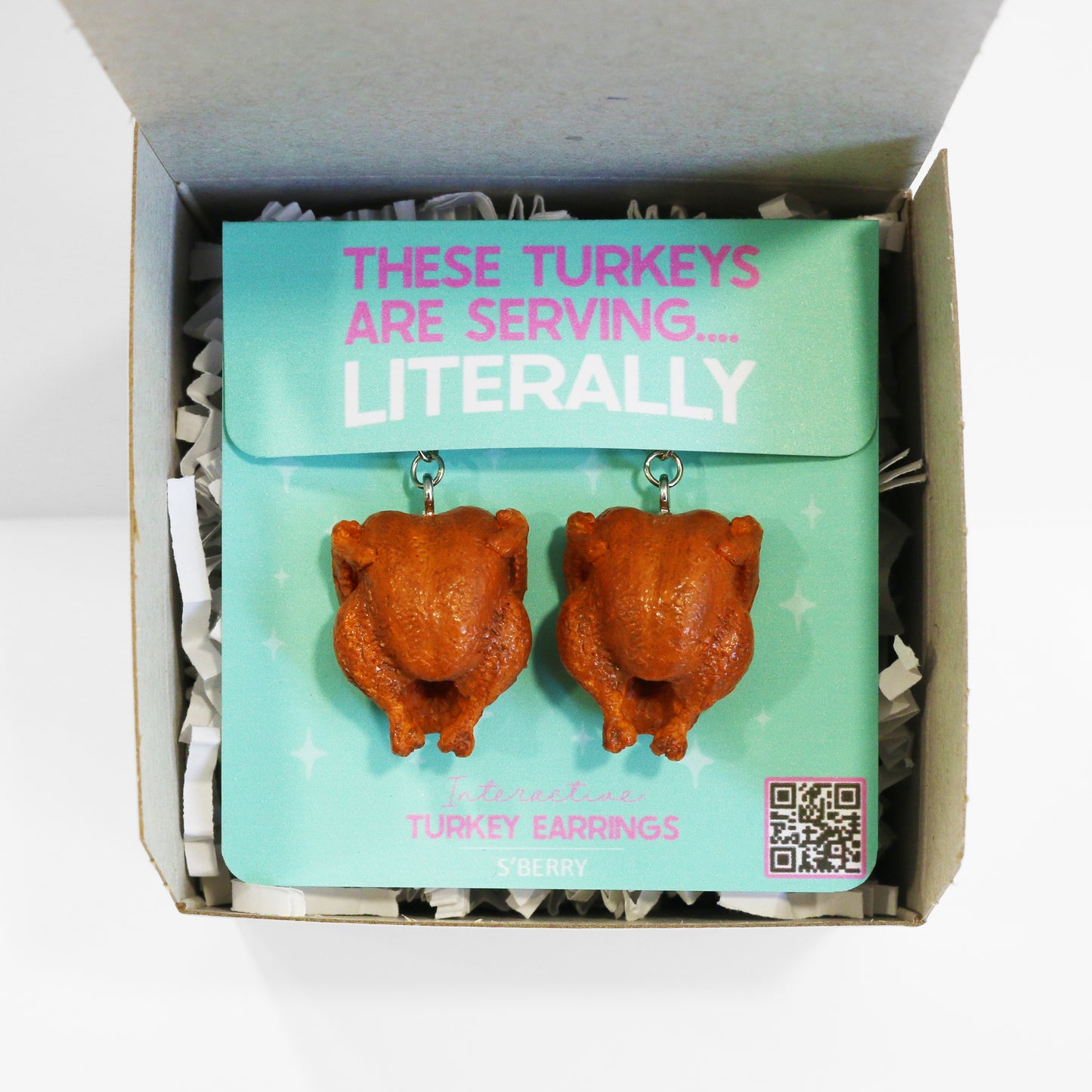 A pair of turkey-shaped earrings displayed on a turquoise card in a gift box with the text "THESE TURKEYS ARE SERVING... LITERALLY" and a QR code at the bottom right.