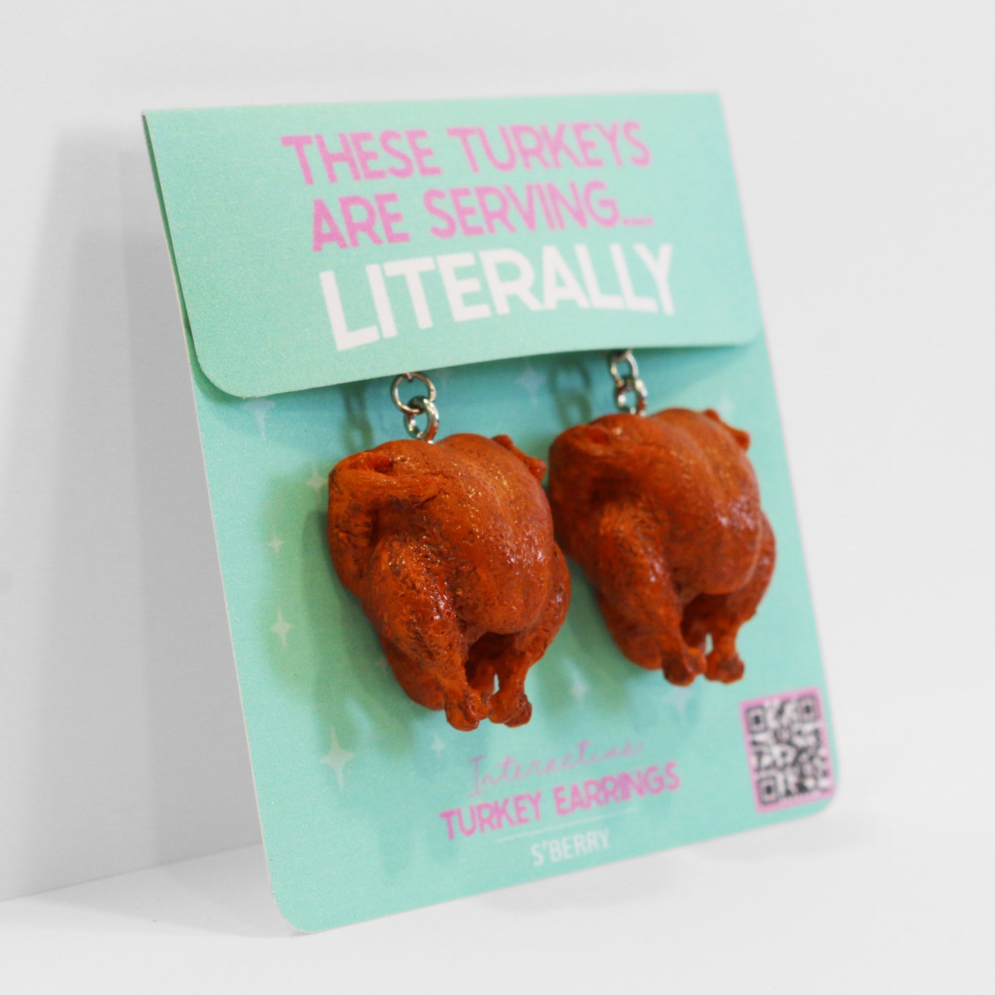 A pair of earrings shaped like roasted turkeys displayed on a turquoise card labeled "These Turkeys Are Serving... Literally.