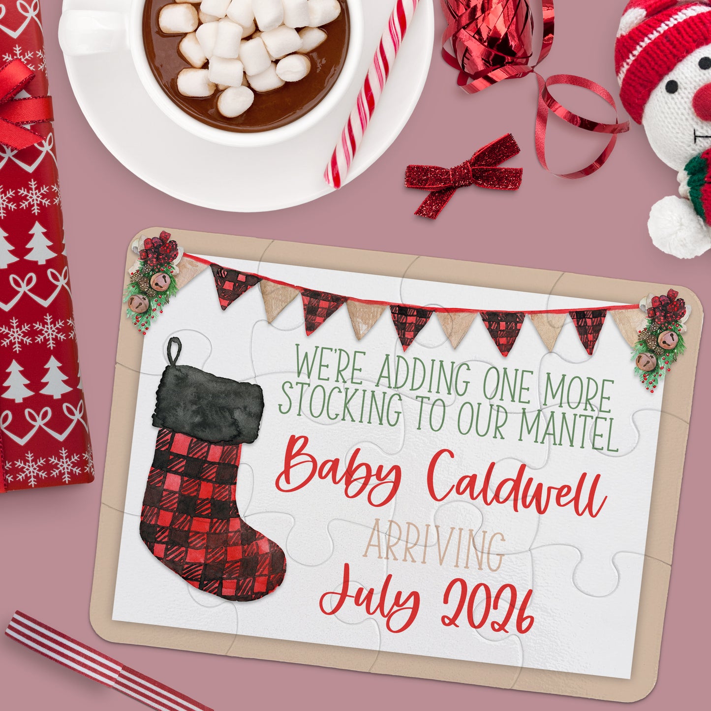 A holiday-themed flat lay with hot chocolate, a snowman, candy cane, and a puzzle announcing "Baby Caldwell arriving July 2026" with a red plaid stocking and festive decor.