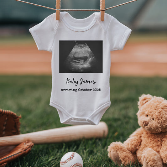 White baby onesie with a sonogram image and text "Baby James arriving October 2025" hangs on a line above a baseball field, next to a teddy bear, bat, and glove.