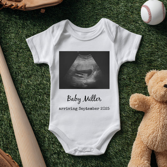 A white baby onesie with an ultrasound image and the text "Baby Miller arriving September 2025" is laid on grass next to a bat, glove, baseball, and teddy bear.