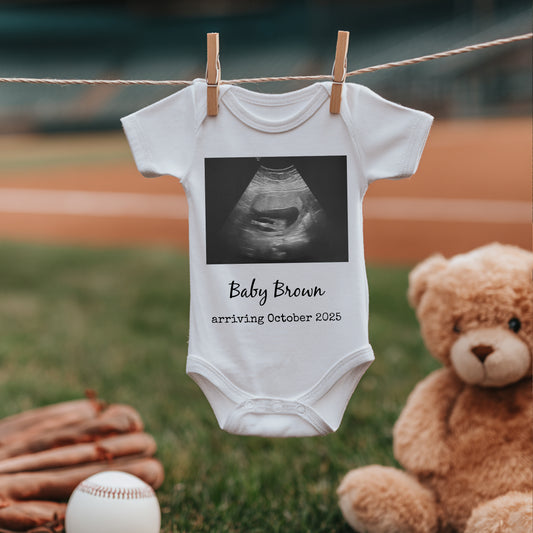 A white baby onesie with a sonogram image and the text "Baby Brown arriving October 2025" hangs on a clothesline, with a teddy bear, baseball glove, and ball nearby.