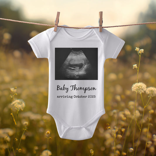 White baby onesie hanging on a clothesline outdoors, featuring a sonogram image and the text "Baby Thompson arriving October 2025.