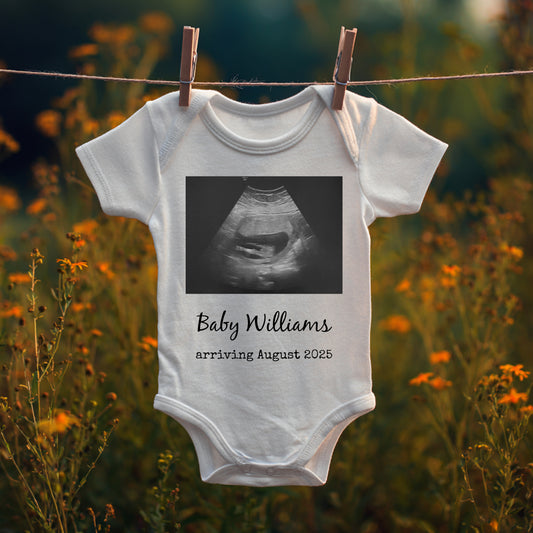 White baby onesie hangs on a clothesline outdoors, displaying a sonogram image with text: "Baby Williams arriving August 2025.