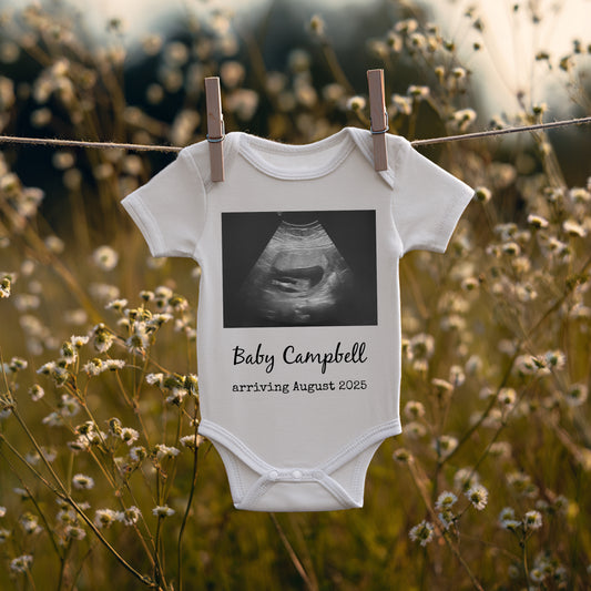 A baby onesie clipped to a clothesline outdoors, displaying a sonogram image and the text “Baby Campbell arriving August 2025.”.