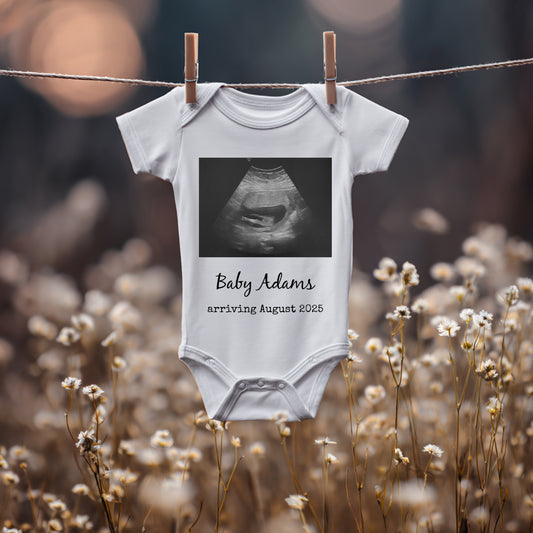 A white baby onesie with a printed ultrasound image and the text "Baby Adams arriving August 2025" hangs on a clothesline above wildflowers.