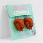 A pair of earrings shaped like roasted turkeys displayed on a turquoise card labeled "These Turkeys Are Serving... Literally.